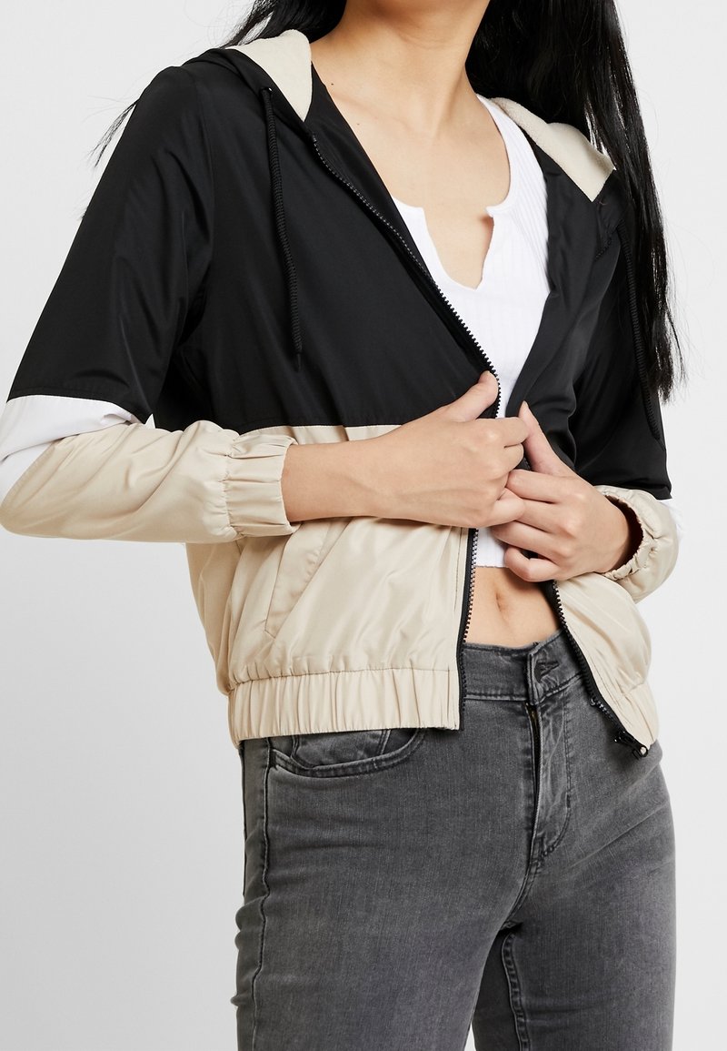 Czarna i beżowa kurtka z kapturem, zapinana na suwak, z elastycznymi mankietami i krótkim krojem. Gładka faktura z panelami w blokach kolorystycznych.