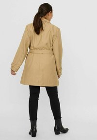 Beige Mantel mit Gürtel und taillierter Silhouette, langen Ärmeln und Manschettendetails, kombiniert mit schwarzen engen Hosen und Stiefeletten.