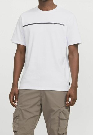 Homme portant un T-shirt blanc à manches courtes avec une ligne noire horizontale et un texte sur la poitrine, associé à un pantalon cargo beige.