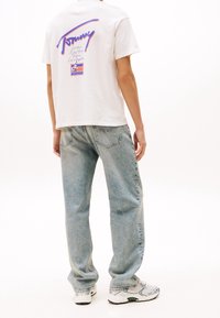 Osoba w białej koszulce z nadrukiem nazw miast i jasnoniebieskich luźnych dżinsach stoi tyłem do kamery na białym tle.