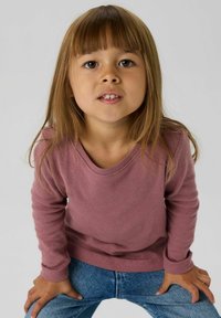 Różowy sweter z długimi rękawami o fakturowanej strukturze i okrągłym dekolcie, zestawiony z niebieskimi jeansami. Dziecko ma długie włosy i neutralny wyraz twarzy.