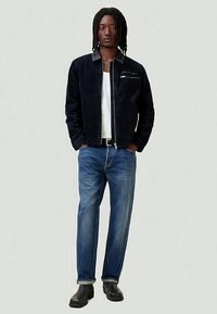 Hombre con rastas, vistiendo una chaqueta negra, camisa blanca, jeans azules y botas negras, de pie frente a un fondo claro y sencillo.