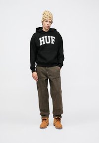 Felpa nera con logo "HUF", pantaloni verde scuro e stivali color sabbia. La persona indossa un berretto color sabbia con pois marroni. Sfondo semplice.