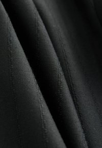 Schwarzer Stoff mit vertikalen Streifen, der eine glatte Textur und subtile Nähdetails aufweist. Das Material wirkt strapazierfähig und strukturiert.