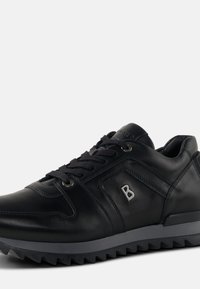 Black leather sneaker with round toe, textured accents, and lace-up closure. Features a gray rubber sole with zigzag tread pattern.