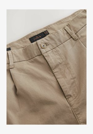 Beige bomuldsbukser med flad front, knaplukning og bæltestropper. Har tonale syninger og en sort mærke med logo.