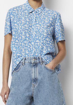 Persoon die een blauw overhemd met korte mouwen en witte bloemenprint draagt, ingestopt in lichtblauwe high-waisted spijkerbroek.