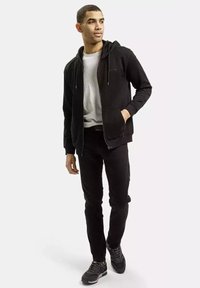 Czarna BD zipper hoodie z przednimi kieszeniami, noszona na jasnoszarym t-shircie. Wąskie czarne spodnie i ciemne buty sportowe dopełniają strój.