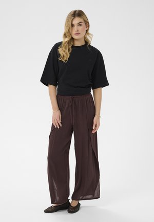 Młoda kobieta o blond włosach, ubrana w czarną bluzkę z krótkim rękawem, luźne brązowe spodnie ze sznurkiem i czarne plecione baleriny, stojąca na białym tle.