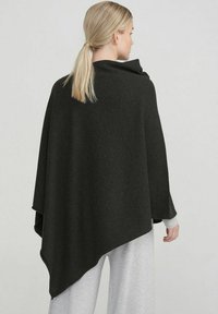 Poncho gris foncé avec un design drapé, texture douce et ourlet asymétrique, porté sur une tenue gris clair, vu de l'arrière.