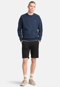 Felpa girocollo blu marina con maniche raglan, abbinata a pantaloni corti neri. Presenta un logo sul petto e calzini chiari con strisce.