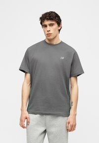 Νέος άνδρας με σκούρα σγουρά μαλλιά, φόρεμα άνετο γκρι μπλουζάκι New Balance και ανοιχτό γκρι φόρμα, στέκεται μπροστά σε απλό φόντο.