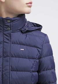 Osoba v námořnickém modrém prošívaném kabátu s vysokým límcem a patentkami, ukazující horní část trupu a krku.