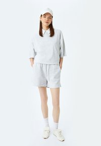 Jongeman in een lichtgrijze oversized sweatshirt, bijpassende short, witte pet, sokken en witte sneakers, staand met haar handen in de zakken.