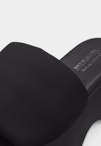 Μαύρο σανδάλι χωρίς λουράκια με υφασμάτινο φαρδύ λουρί και μαλακή σόλα, κοντινή λήψη που δείχνει τη λεπτομέρεια του υφάσματος και το ανάγλυφο κείμενο της μάρκας στην εσωτερική σόλα.