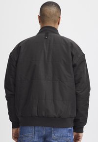 Svart quiltet jakke med ribbet krage og mansjetter, med en jevn tekstur og en logoferd på baksiden, parret med blå jeans.