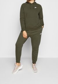 Sudadera Nike de color verde oliva con un bolsillo frontal y logo, combinada con pantalones jogger a juego. El modelo lleva zapatillas Nike de color rosa claro con suelas blancas.