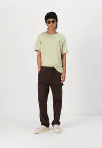 Jauns vīrietis, tērpies gaiši zaļā t-kreklā, tumši brūnos šortos ar aukliņām, bēšos kedās un ar apaļajām saulesbrillēm, stāv uz balta fona.