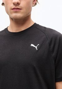 Νεαρός άνδρας που φοράει μαύρο μπλουζάκι Puma με λευκό λογότυπο Puma στο αριστερό μέρος του στήθους, σε απλό λευκό φόντο.