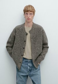 Brauner, strukturierter Cardigan mit rundem Ausschnitt und Knopfverschluss, getragen über einem beigen Hemd und blauen Jeans, vor einem einfarbigen Hintergrund.