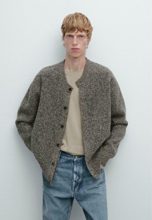 Cardigan marrone con texture, con scollatura rotonda e chiusura a bottoni, indossato sopra una maglietta beige e jeans blu, su uno sfondo semplice.
