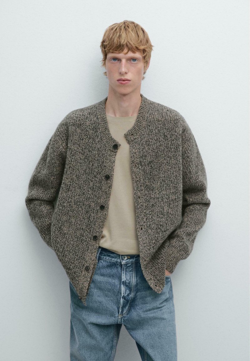 Brauner, strukturierter Cardigan mit rundem Ausschnitt und Knopfverschluss, getragen über einem beigen Hemd und blauen Jeans, vor einem einfarbigen Hintergrund.