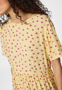 Γυναίκα με καστανά σγουρά μαλλιά που φοράει κίτρινο φόρεμα με κοντά μανίκια και ροζ λουλουδάτο σχέδιο σε ανοιχτό φόντο.