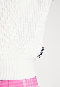 Maglione bianco a maglia con texture a coste e etichetta nera "HUGO" sul lato, abbinato a una gonna a quadretti rosa. Il maglione ha una vita aderente.