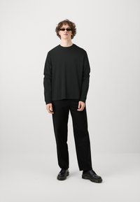 Zwart langauw trui met een relaxte pasvorm, gecombineerd met rechte zwarte broek en zwarte veterschoenen. Model draagt slanke zonnebril.