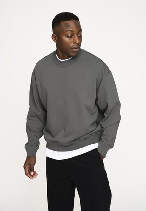 Grå sweatshirt med ribbet hals og manchetter, der har en afslappet pasform og sidesømme. Båret over en hvid skjorte og parret med sorte bukser.