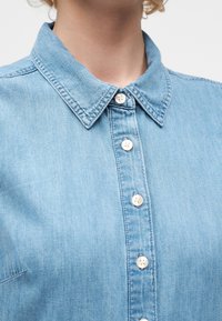 Κοντινό πλάνο ατόμου που φοράει ανοιχτό μπλε πουκάμισο από τζιν με μυτερό γιακά και λευκά κουμπιά μπροστά.