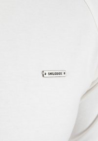 Weißes T-Shirt aus Stoff mit einem kleinen silbernen rechteckigen Etikett, auf dem der Text "SMILODOX" steht. Das Shirt hat eine glatte Textur.