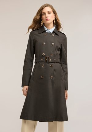 Frau trägt einen schwarzen, doppelt geknöpften Trenchcoat mit Gürtel, langen Ärmeln und beigen Hosen vor einem einfarbigen beigen Hintergrund.