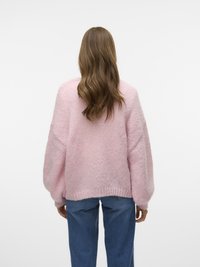 Donna con capelli castani ondulati che indossa un maglione morbido rosa chiaro e jeans blu, in piedi di fronte a uno sfondo bianco uniforme.