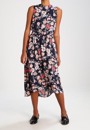 Ærmeløs navy midi-kjole med pink og hvidt blomsterprint, indsnøret talje, parret med sorte skinnende loafers på en stående model.
