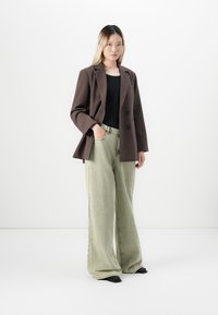 Jeune femme portant un blazer marron, un haut noir, un jean beige à jambes larges et des chaussures noires, se tenant devant un fond blanc.