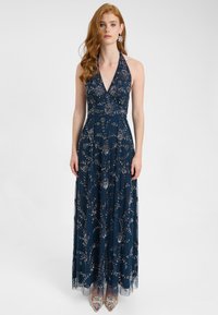 Marineblå halterneck-gown med intrikate perle- og paletmønstre i blomstermønster, lagdelt tylskørt, der når gulvet, parret med klare dekorerede stilethæle.