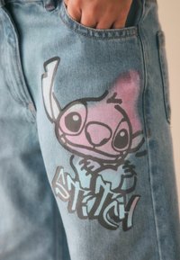 Blugi din denim cu un personaj de desen animat colorat și cuvântul „skitch” imprimat pe coapsă, mână parțial în buzunar.