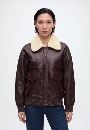 Femme portant une veste en cuir marron foncé avec un grand col en shearling crème et des poches avant à rabat, debout devant un fond blanc uni.