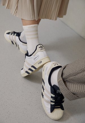 Deux personnes portant des baskets Adidas Superstar blanches avec des bandes noires, l'une en jupe plissée et chaussettes, l'autre en pantalon à carreaux.