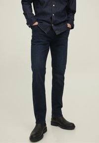 Man i mörkblå denimjeans, en matchande mörkblå skjorta med knappar och svarta läderstövlar, står med händerna i fickorna.