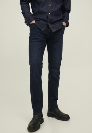 Hombre vestido con jeans de mezclilla azul oscuro, una camisa abotonada azul oscuro a juego y botas de cuero negras, de pie con las manos en los bolsillos.