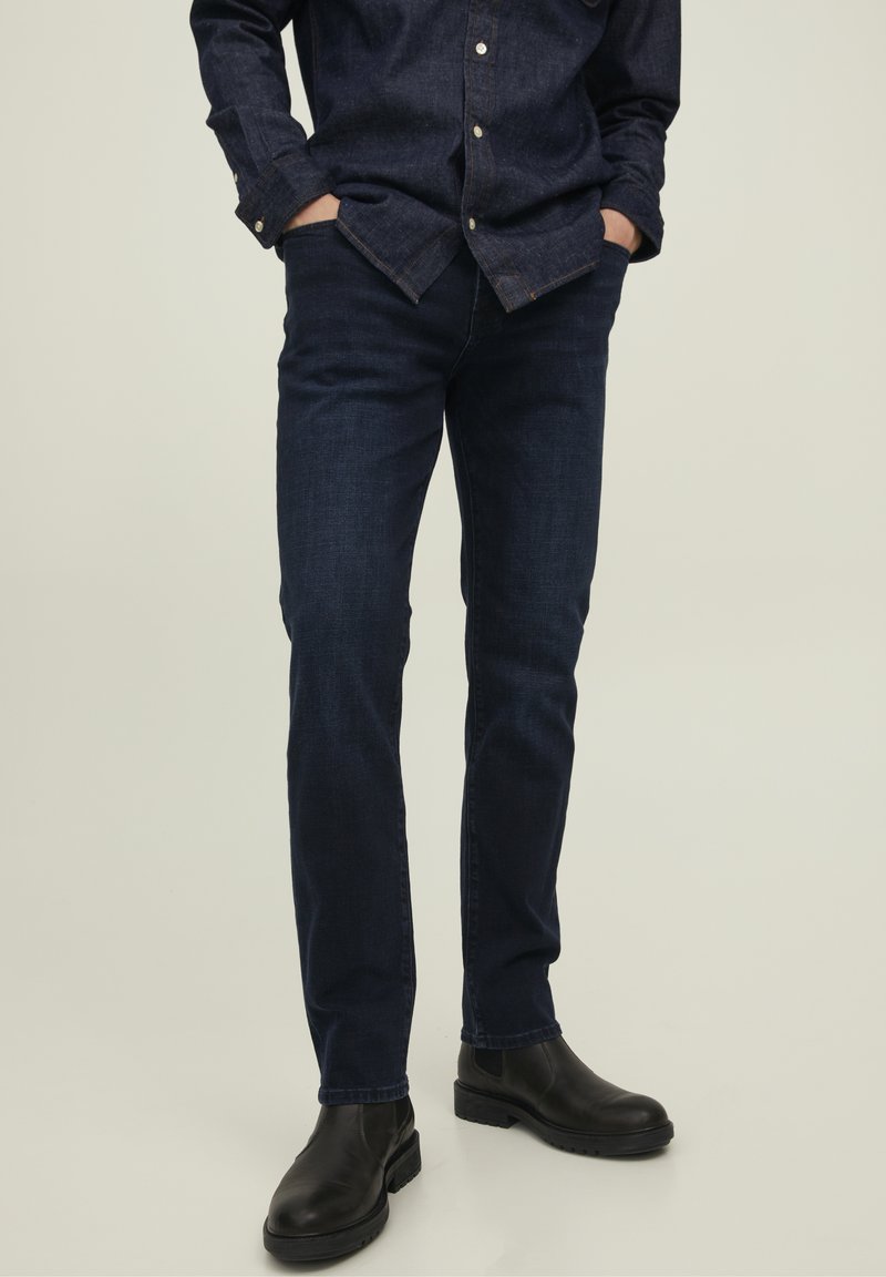 Man i mörkblå denimjeans, en matchande mörkblå skjorta med knappar och svarta läderstövlar, står med händerna i fickorna.