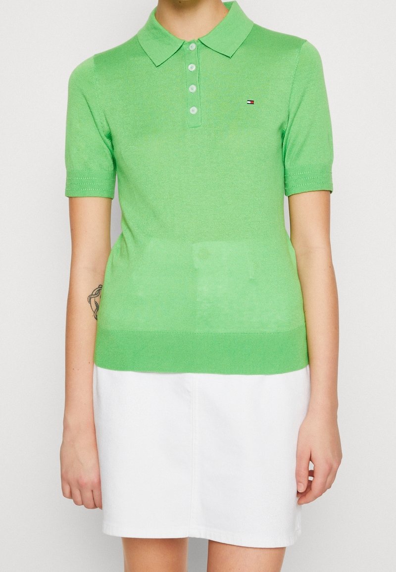 Personne portant un polo à manches courtes vert clair avec quatre boutons et un petit logo sur la poitrine, associé à une jupe blanche.