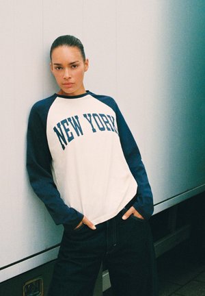 Jeune femme aux cheveux foncés attachés porte un t-shirt à manches longues blanc et marine avec le texte "NEW YORK", debout les mains dans les poches.