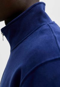 Collo a zip blu navy realizzato con un tessuto morbido e texture. Il colletto è alto, evidenziando i dettagli delle cuciture e una silhouette pulita.