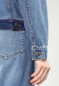 Light blue denim jacket featuring a button closure, contrasting patch, and visible stitching. Cuffs have metal buttons and a frayed edge.