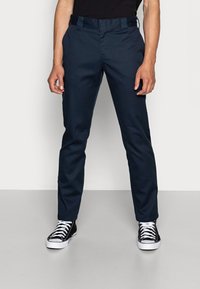 Homme portant un pantalon slim bleu marine et des baskets montantes noires avec des lacets blancs, debout devant un fond clair uni.