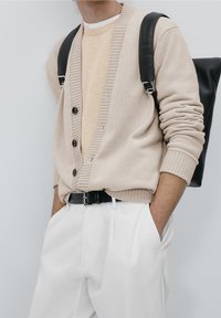 Cardigan beige tricoté avec une texture côtelée, cinq boutons et un col en V sur un pull beige clair ; associé à un pantalon blanc coupé sur mesure et une ceinture noire.