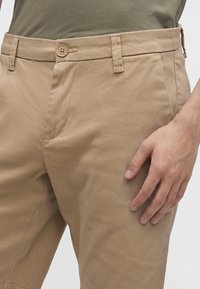 Beige trousers with button and belt loops worn with olive green shirt, hand resting on thigh, against neutral background.